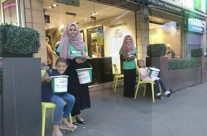 Two women, stood, and two children, sat on chairs, pose in front of shop with buckets.